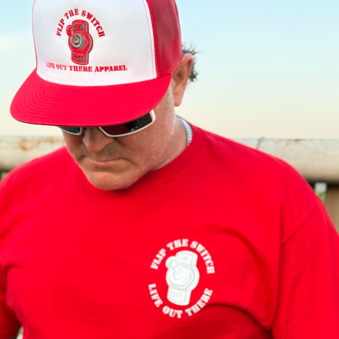 A person wearing a red Unisex - Flip the Switch Tee by Life Out There Apparel and a red and white trucker cap gazes downward against a clear sky backdrop with an indistinct object, embodying the adventurous spirit of their anniversary launch.