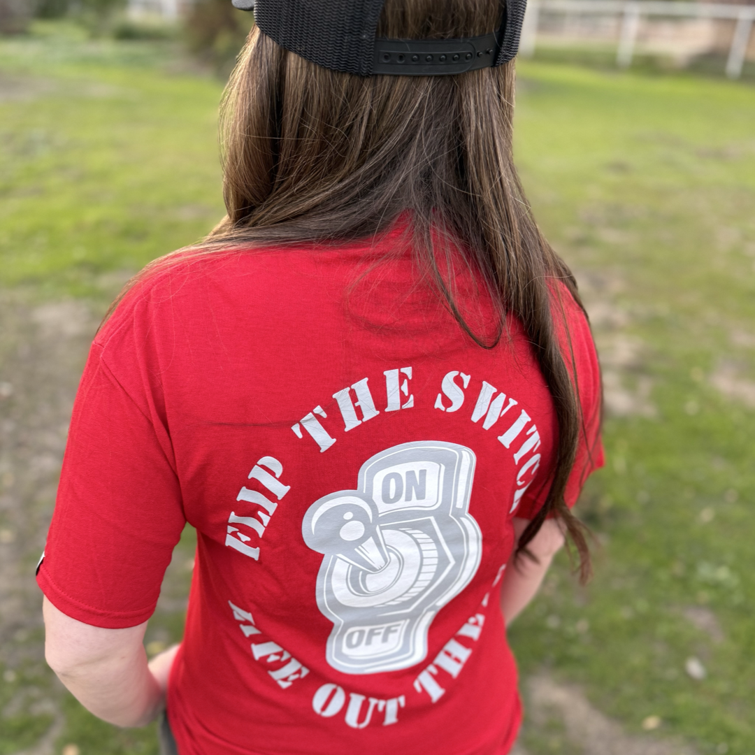 A person with long hair, wearing Life Out There Apparels Unisex Flip the Switch Tee in red and a black cap, stands on grass. The shirt displays a light switch labeled ON and OFF with the words FLIP THE SWITCH, TAKE OUT THE... encircling it.