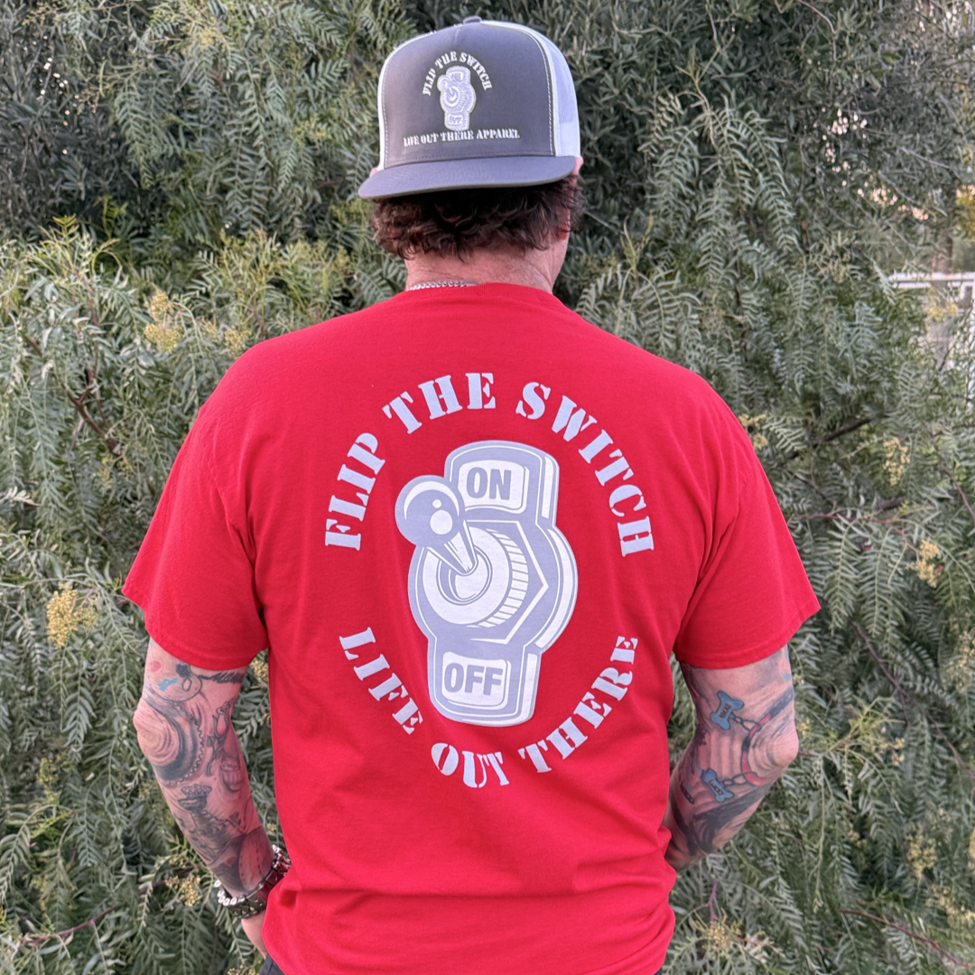 A man wears Life Out There Apparels Unisex Flip the Switch Tee in red, featuring a bold slogan and switch graphic. He pairs it with a black cap with a patch, standing amongst greenery, displaying his tattooed arms during the anniversary launch.