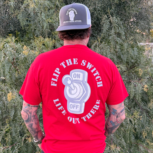 A tattooed person stands outdoors, back to the camera, wearing the red Flip the Switch Tee from Life Out There Apparel to celebrate its anniversary launch.