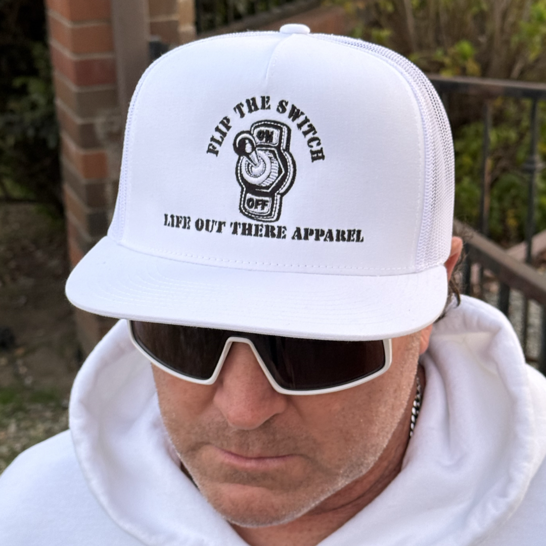 A person in a FLIP THE SWITCH - Flat Bill SnapBack Trucker Hat by Life Out There Apparel announces its anniversary launch. They complete the look with large white sunglasses and a hoodie while standing outdoors near a brick wall.