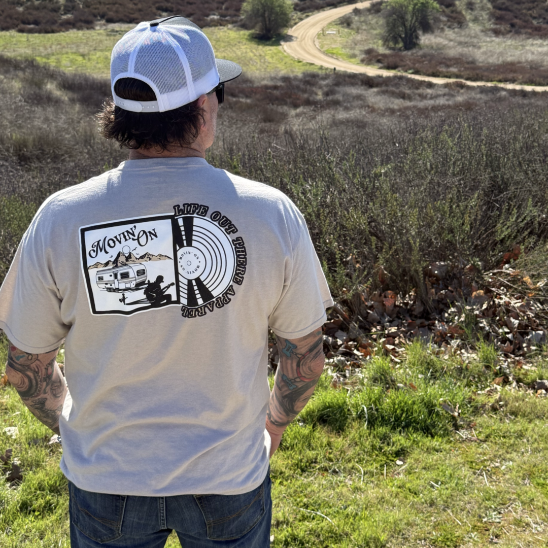 Wearing the Life Out There Apparel Unisex Movin On Tee in Sandstone, a person in a cap and sunglasses stands on grassy terrain, facing away as a dirt road winds behind them. The shirt features a camper, a man playing guitar, and MOVIN ON text.