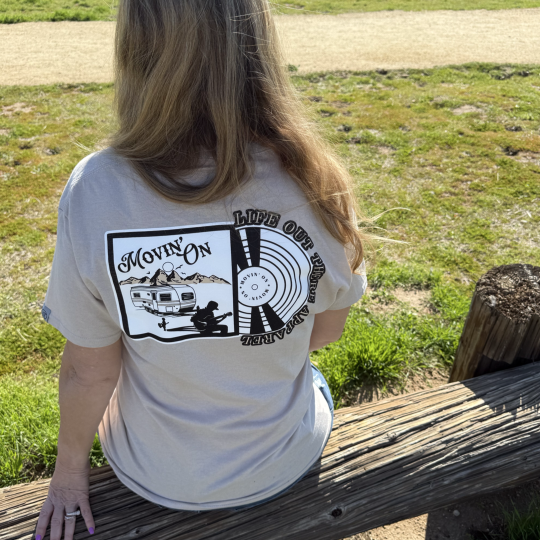 A woman with long hair sits on a wooden bench outdoors, wearing the Life Out There Apparel Unisex Movin On Tee in Sandstone, which features camper and record graphics with the text “Movin’ On” and "Life Out There Apparel".