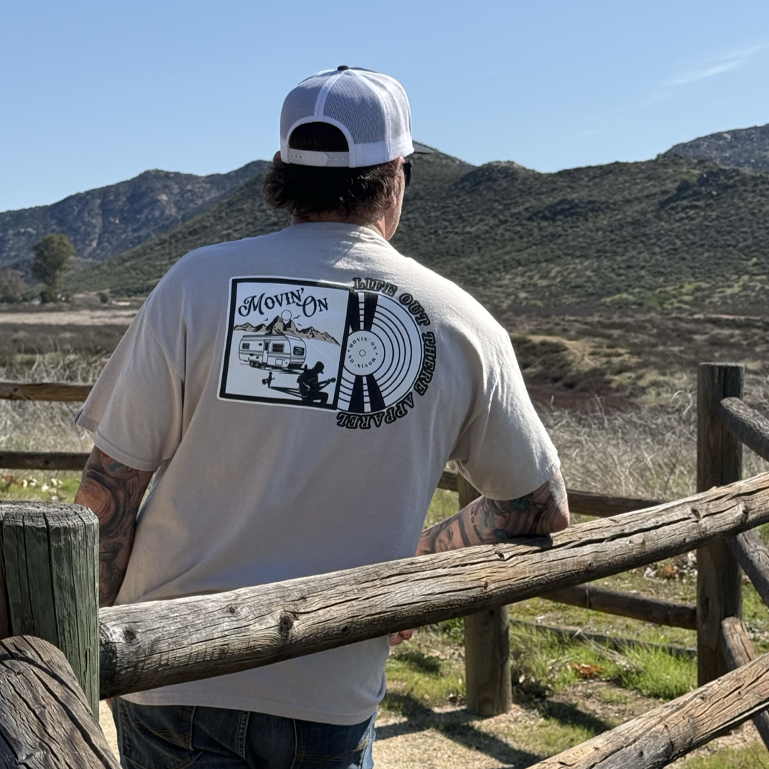 A man in a white cap and the Life Out There Apparel Unisex Movin’ On Tee in Sandstone stands by a wooden fence, gazing at sunny hills—perfectly capturing that MOVIN’ ON spirit.