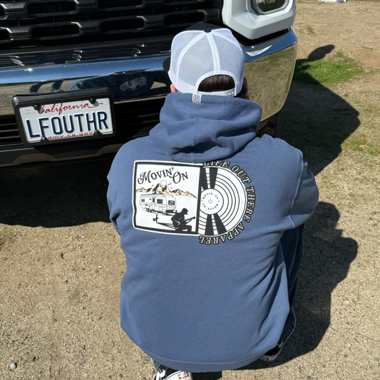 A person in a white cap crouches near a truck with a California license plate, wearing the Movin' On Hoodie - Denim Blue by Life Out There Apparel, featuring a camper, mountains, and "Movin On" text inspired by country music on the back.