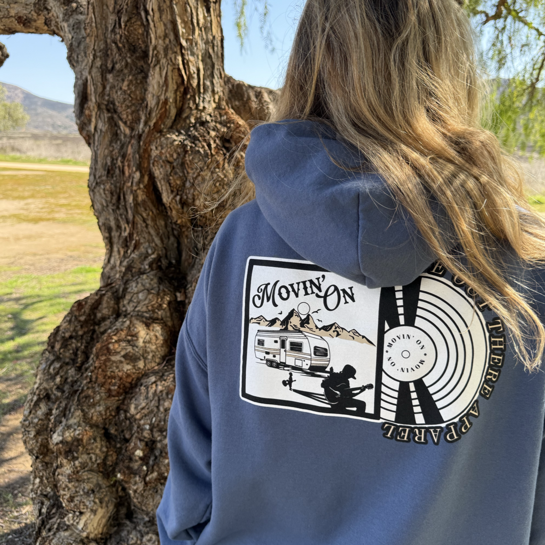 Someone in a Life Out There Apparel Unisex Movin On Hoodie in Denim Blue, featuring Jeremiah Watsons camper, mountains, and guitar graphic, stands by a tree in the sun.