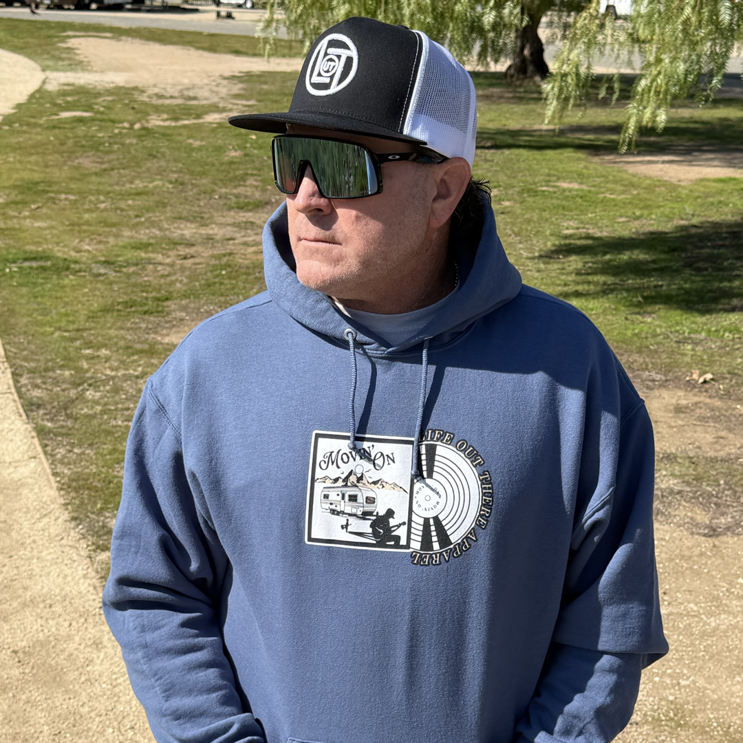 Wearing the Unisex - Movin On Hoodie in Denim Blue by Life Out There Apparel, a man stands outside on a sunny day, sunglasses and cap on, gazing to the side among grass, a dirt path, and trees—embracing that MOVIN ON spirit.