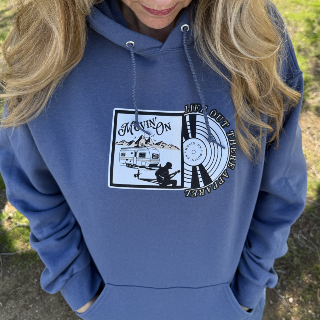 A woman with long blonde hair wears the Unisex - Movin On Hoodie in Denim Blue by Life Out There Apparel, featuring a camper, mountains, a person by a fire, and the text “MOVIN’ ON,” design by "Life Out There Apparel".