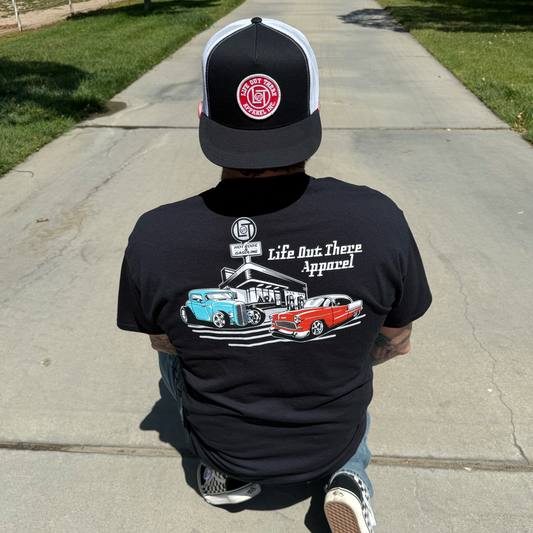 A person in the Cruzin' Tee - Black by Life Out There Apparel, featuring a hot rod graphic, kneels on a sidewalk bordered by grass—making this shirt a perfect Father’s Day gift.