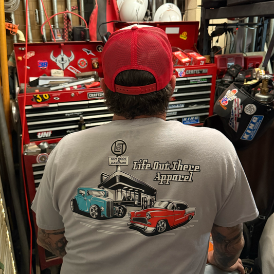 A man in a red cap and gray “Cruzin' Tee - Silver” by Life Out There Apparel, featuring vintage cars like a '55 Chevy Bel Air, sits in a garage with red toolboxes—an ideal Father's Day gift for car lovers.