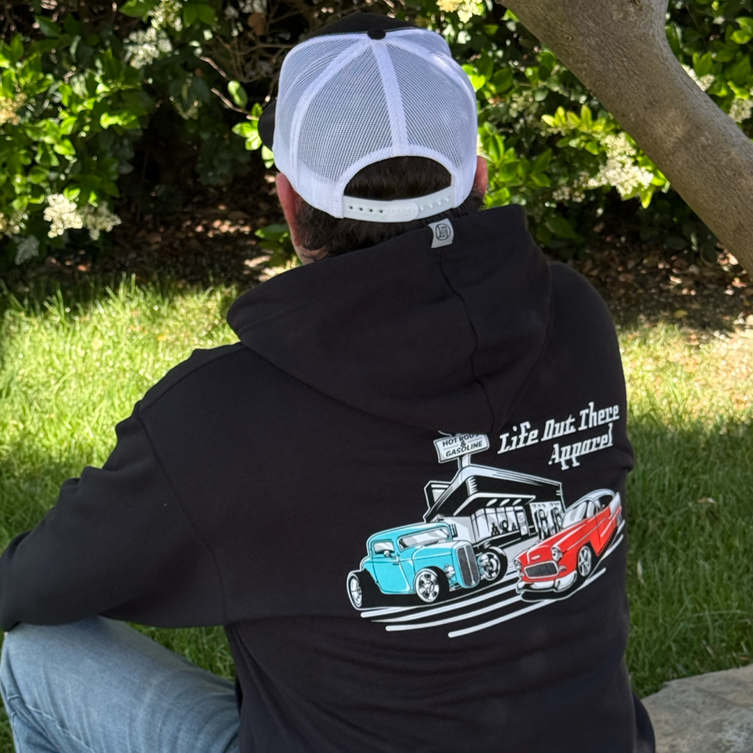 Wearing the Unisex Cruzin Zip-Up Hoodie in black by Life Out There Apparel, featuring two vintage cars including a classic Chevy Bel Air, and a white mesh cap, someone sits on grass—an ideal Fathers Day gift for hot rod fans.