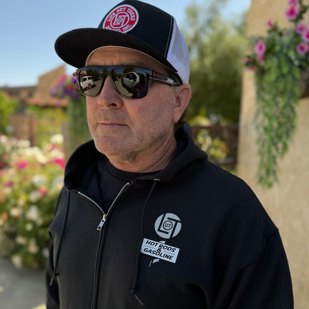 A man enjoys the sun in a Life Out There Apparel Unisex Cruzin Zip-Up Hoodie—Black, sunglasses, and a black-and-white cap, standing near blooming plants and a beige building—an ideal Fathers Day vibe.