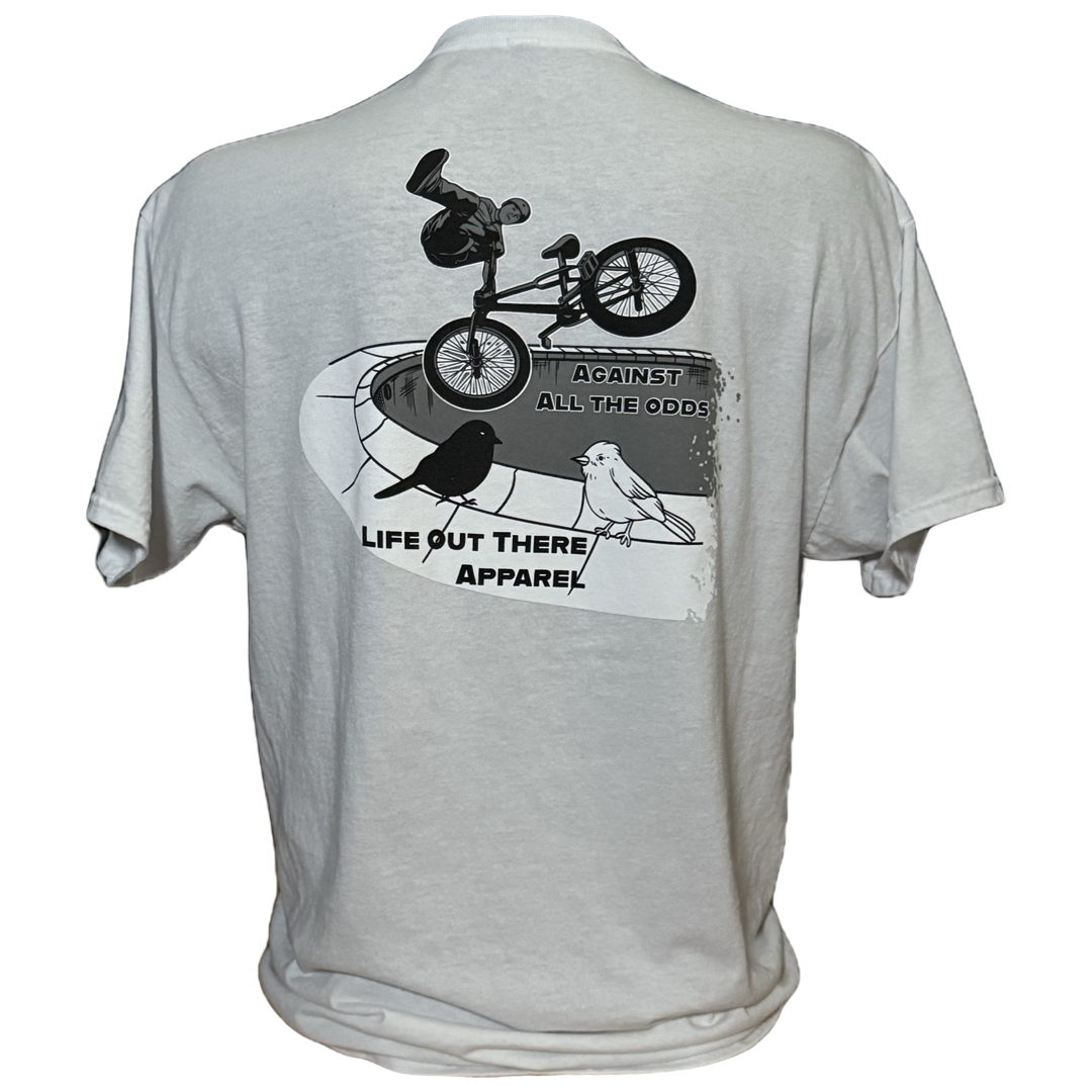 The Unisex Against All The Odds / The Ride Tee in white by Life Out There Apparel features BMX legend John Buultjens performing a skate park trick with a bird and the text AGAINST ALL THE ODDS.