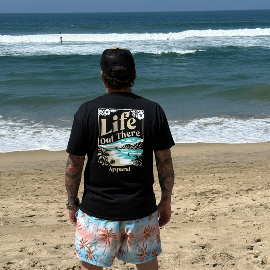 On a sandy North Shore Oahu beach, a person faces the ocean in colorful swim trunks and The Aloha Tee - Black by Life Out There Apparel. Waves roll in behind them, radiating true Hawaiian vibes.