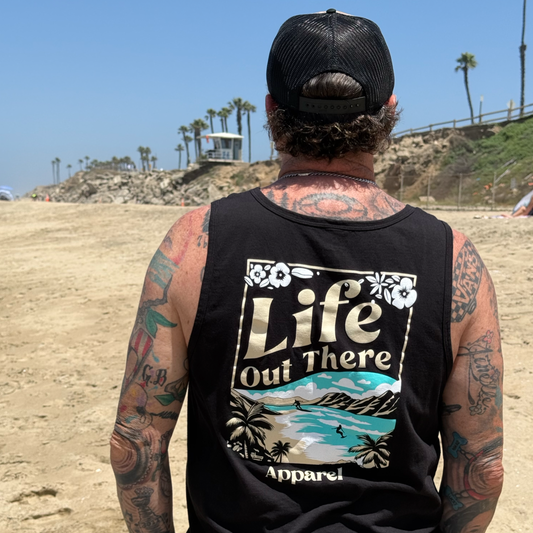 A tattooed man in a black cap and The Aloha Tank Top by Life Out There Apparel stands on a sandy North Shore Oahu beach, facing the ocean; the shirt’s tropical design captures true Hawaiian vibes.
