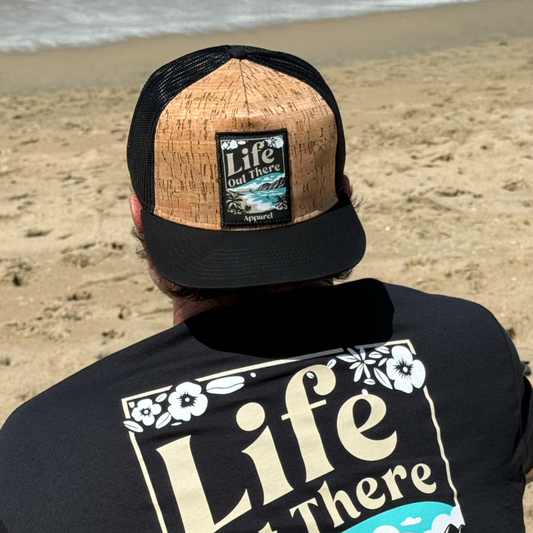 Facing the ocean on a sandy North Shore beach, someone wears the ALOHA - Flat Bill SnapBack Trucker Hat in Cork/Black by Life Out There Apparel, paired with a floral Life Out There shirt for authentic Hawaiian vibes.