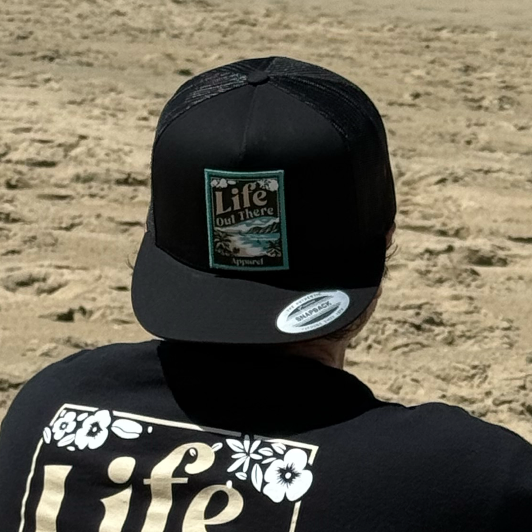 At the beach, someone wears a Life Out There Apparel ALOHA Flat Bill SnapBack Trucker Hat in black and a black shirt with white Life print, capturing Hawaiian vibes with Oahu’s North Shore and sandy beach in the background.