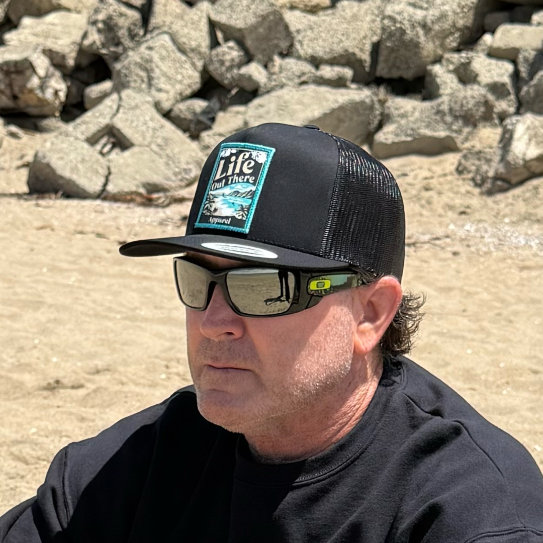 A man in sunglasses, a black shirt, and the Life Out There Apparel ALOHA - Flat Bill SnapBack Trucker Hat (Black) relaxes on a sandy North Shore of Oahu beach with large rocks, taking in the Hawaiian vibes.
