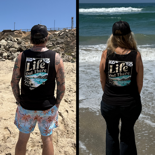 A man and woman stand on the beach in matching Life Out There Apparel’s The Aloha Tank Top - Black, featuring a tropical graphic and “Life Out There” text, with waves and sand in the background.