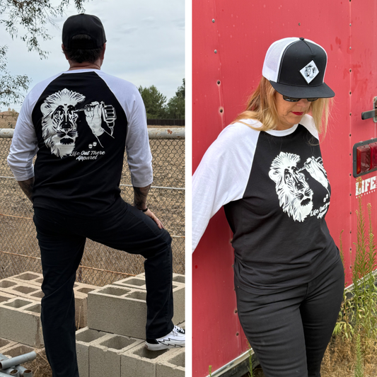 Two people wear matching Life Out There Apparel Unisex - Not All There 3/4 Sleeve Raglan T Shirts in black and white. Each tagless shirt features a lion and fist graphic with text on the back, plus a lion design on the front. They pose by a fence and red wall outdoors.