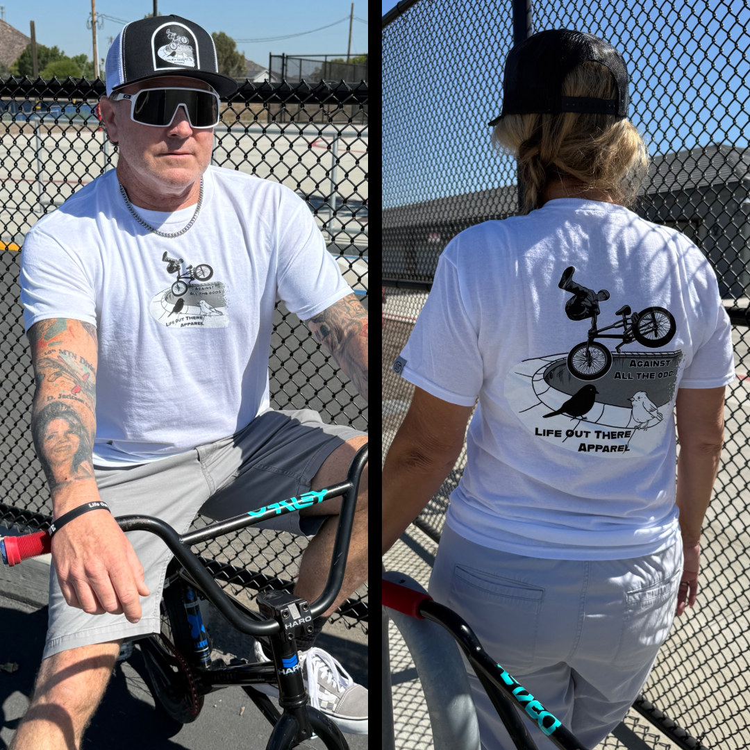 Two people by a black wire fence wear matching Life Out There Apparel Against All The Odds / The Ride unisex white tees, featuring a BMX rider graphic and slogan, paired with grey shorts as they stand next to their bikes.