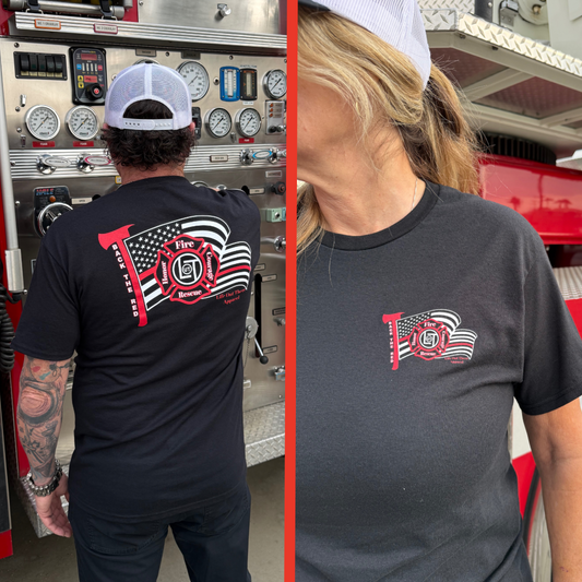 Two people stand near a fire truck in firefighter gear, both wearing the Life Out There Apparel Unisex Back The Red Tee in black, featuring an American flag and Fire Rescue logo. One adjusts the truck controls while the other faces the camera.