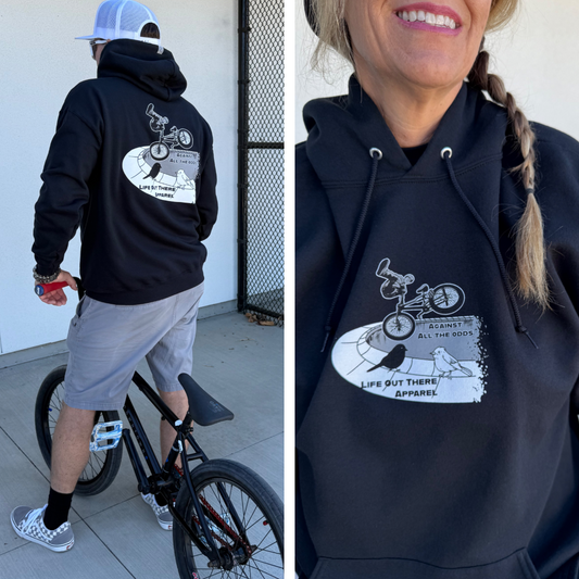 A person in the Unisex - Against All The Odds / The Ride Hoodie - Black by Life Out There Apparel stands near a BMX bike; a close-up reveals the design featuring BMX rider John Buultjens and AGAINST ALL THE ODDS text on the chest.