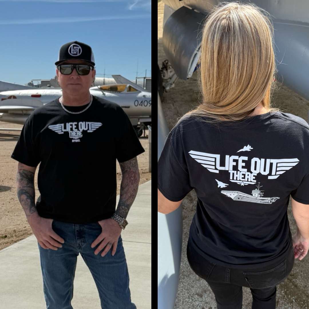 A man and a woman both wear black Unisex The Hard Deck Tee from Life Out There Apparel, featuring aviation-themed graphics inspired by the US Military. The mans shirt is showcased in front of fighter jets, while the womans is seen from the back near an aircraft.