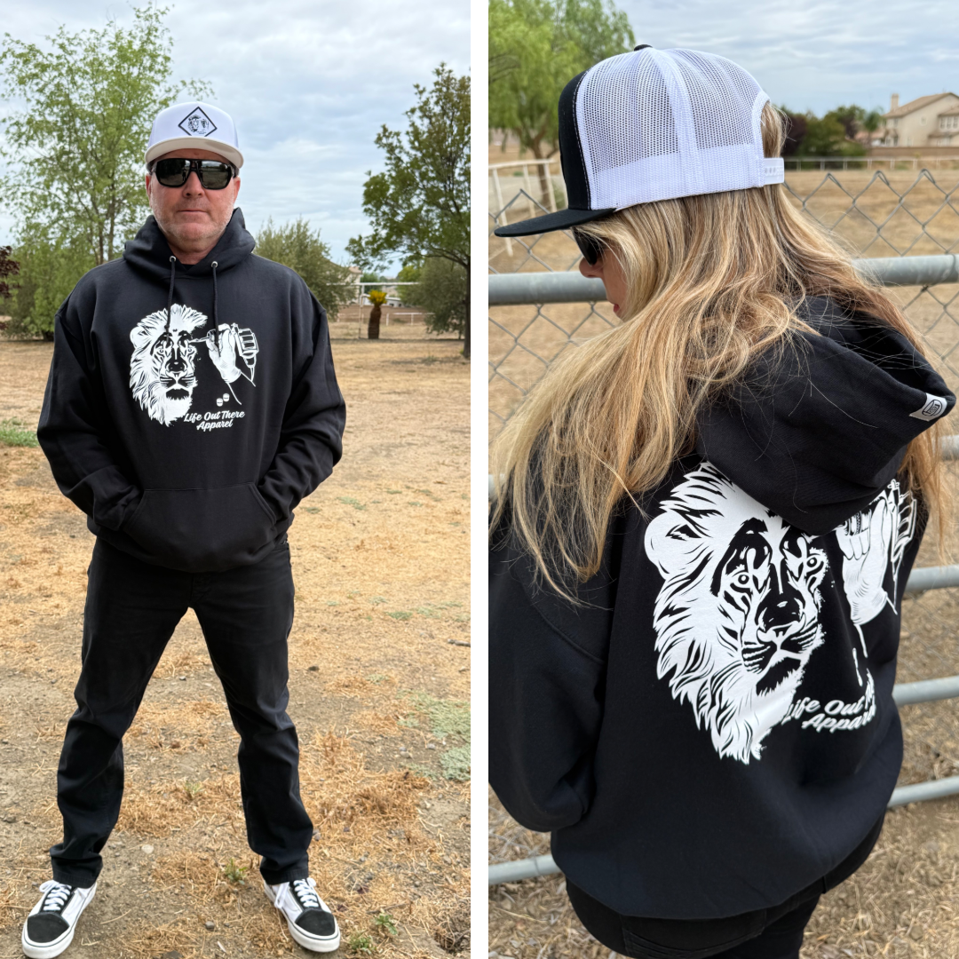 Two people outdoors wear Life Out There Apparel’s Unisex - Not All There Hoodie in black, featuring a white lion graphic and text on the back. Both style the tagless hoodie with hats and sunglasses—one faces forward, one stands sideways by a fence.