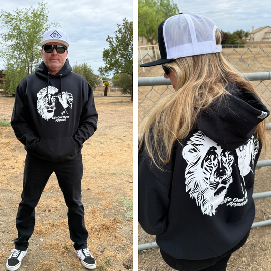 Two people outdoors wear Life Out There Apparel’s Unisex - Not All There Hoodie in black, featuring a white lion graphic and text on the back. Both style the tagless hoodie with hats and sunglasses—one faces forward, one stands sideways by a fence.