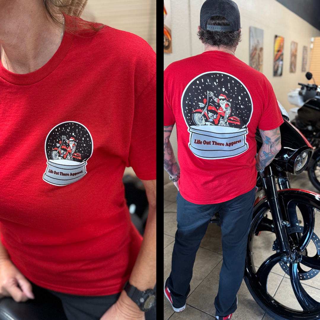 A man and woman wear matching "Unisex - Dashing Through The Snow Tee - Red" by Life Out There Apparel, featuring a snow globe Santa on a motorcycle; the man stands by a black motorcycle indoors.