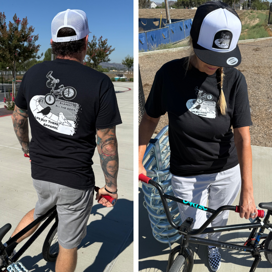 Two people wearing Life Out There Apparel’s Unisex - Against All The Odds / The Ride Tee in black, featuring a graphic of a BMX rider and tributes to “Always in the Loop,” “Live Out the Departure,” and BMX legend John Buultjens, stand with BMX bikes outdoors.