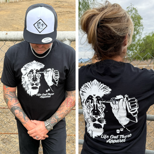 Two people wear the Unisex - Not All There Tee in black by Life Out There Apparel, a soft cotton-poly blend with a lion tattoo graphic and brand text. One faces forward, head down; the other is seen from behind, outdoors near a fence.