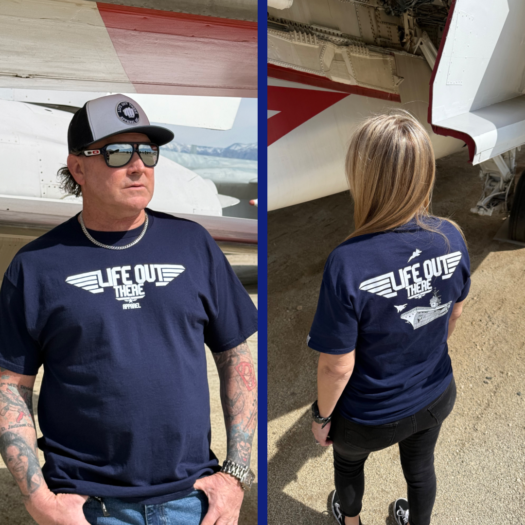 A man and woman in matching navy The Hard Deck Tee by Life Out There Apparel exude daring. The man, with sunglasses and hat, stands strong beside a TOPGUN MAVERICK-style aircraft. From the back, her flowing hair symbolizes readiness for the HARD DECK challenge.