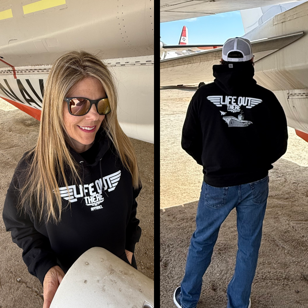 A woman and a man stand by an aircraft with a TOPGUN vibe. The woman wears sunglasses and the Life Out There branded Unisex - The Hard Deck Hoodie in black, while the man, in a cap and matching hoodie, faces away like an iconic US Military fighter jet pilot.