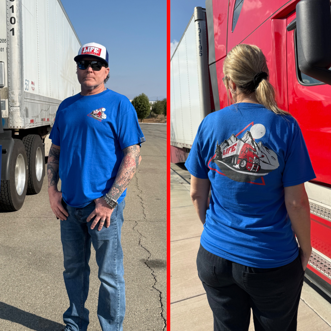 A man and a woman, seemingly truck drivers, stand near a red truck on a sunny day. The man on the left is in a blue shirt and hat. The woman, facing away, wears Life Out There Apparels 10-4 Good Buddy Tee - Royal, featuring 50% cotton with a truck graphic on the back.