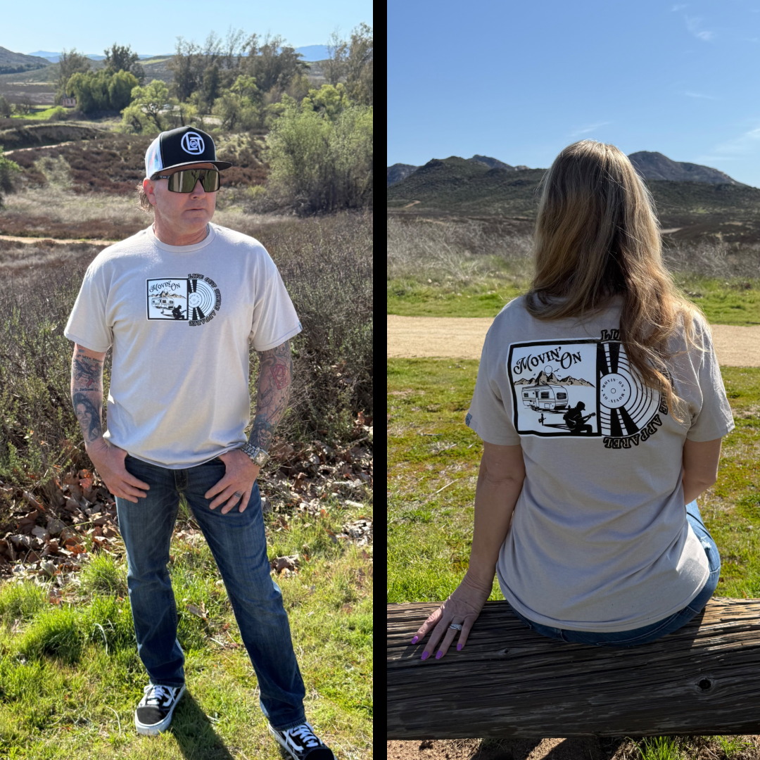 A man wears the Life Out There Apparel Unisex Movin On Tee in Sandstone with jeans, while a woman on a bench shows off its back graphic. Both are outdoors on grassy hills beneath a blue sky.