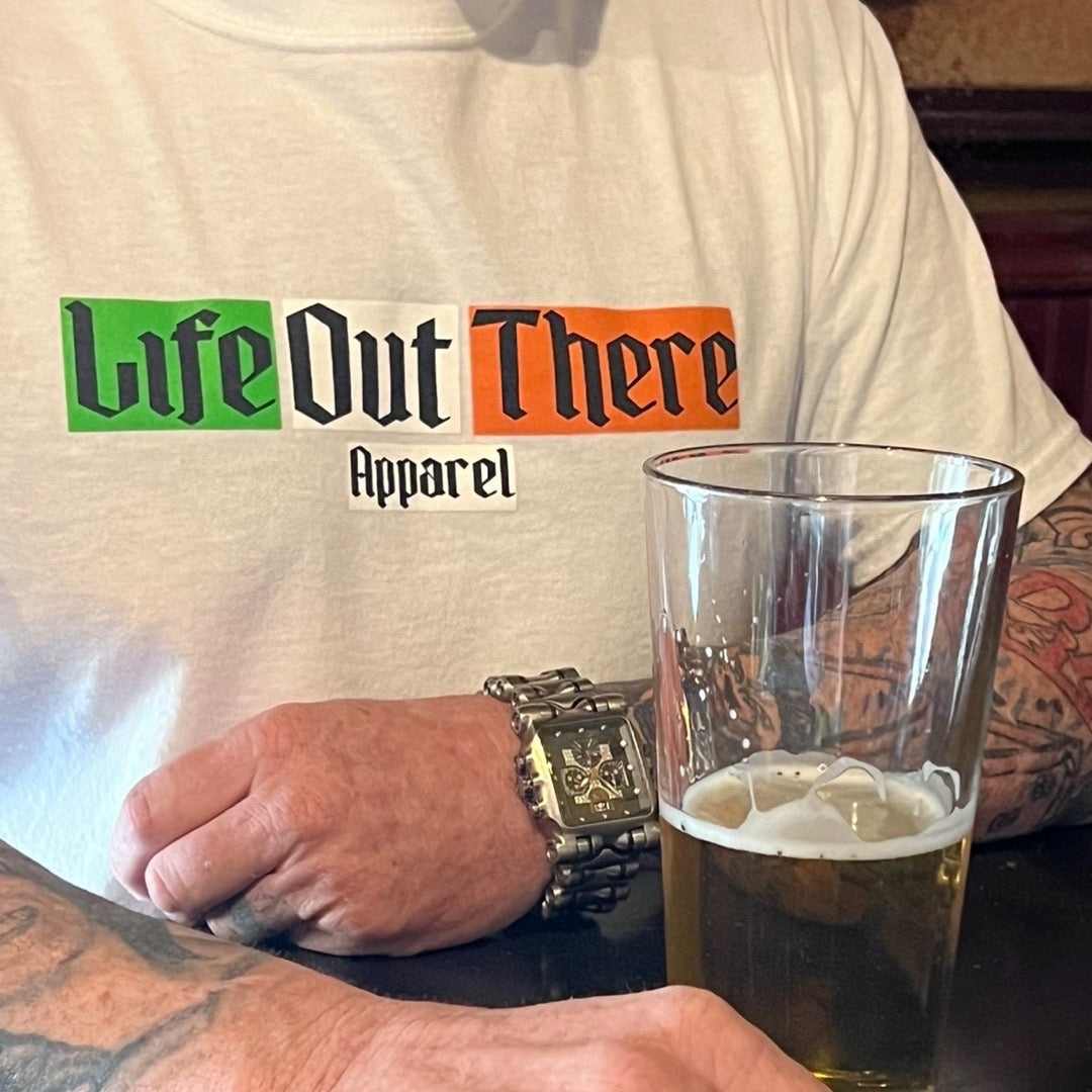 Life Out There Apparel “Capt. Patrick” Unisex tee shirt in white is a collaboration of an American flag with an Irish twist and a leprechaun boat Captain looking on. This is a photo,of a man wearing the shirt in a pub, with a beer.  