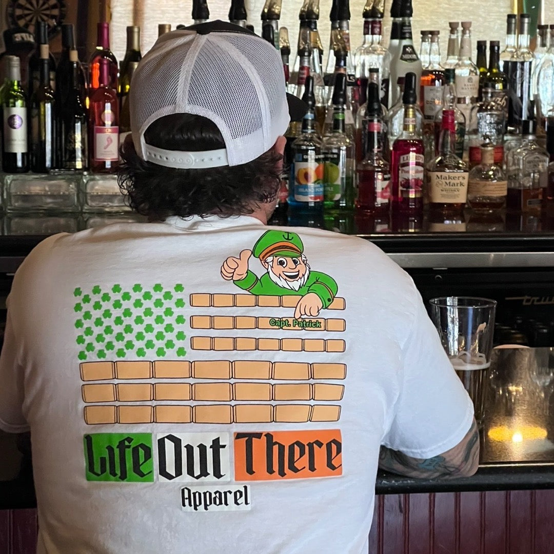 Life Out There Apparel “Capt. Patrick” Unisex tee shirt in white is a collaboration of an American flag with an Irish twist and a leprechaun boat Captain looking on. This is a photo,of a man wearing the shirt at a bar.  
