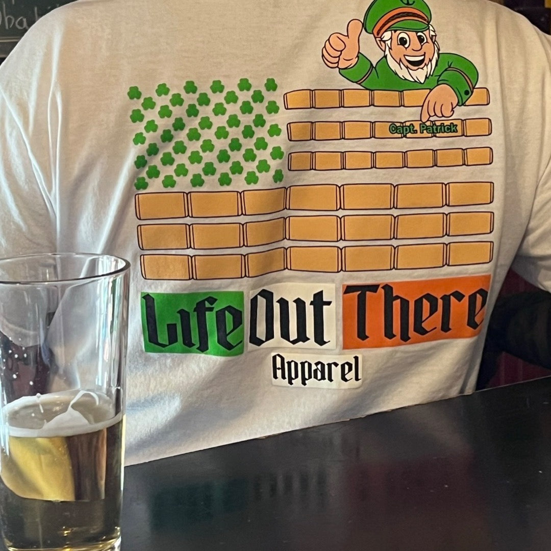Life Out There Apparel “Capt. Patrick” Unisex tee shirt in white is a collaboration of an American flag with an Irish twist and a leprechaun boat Captain looking on. This is a photo,of a man wearing the shirt at a bar shooting darts. 