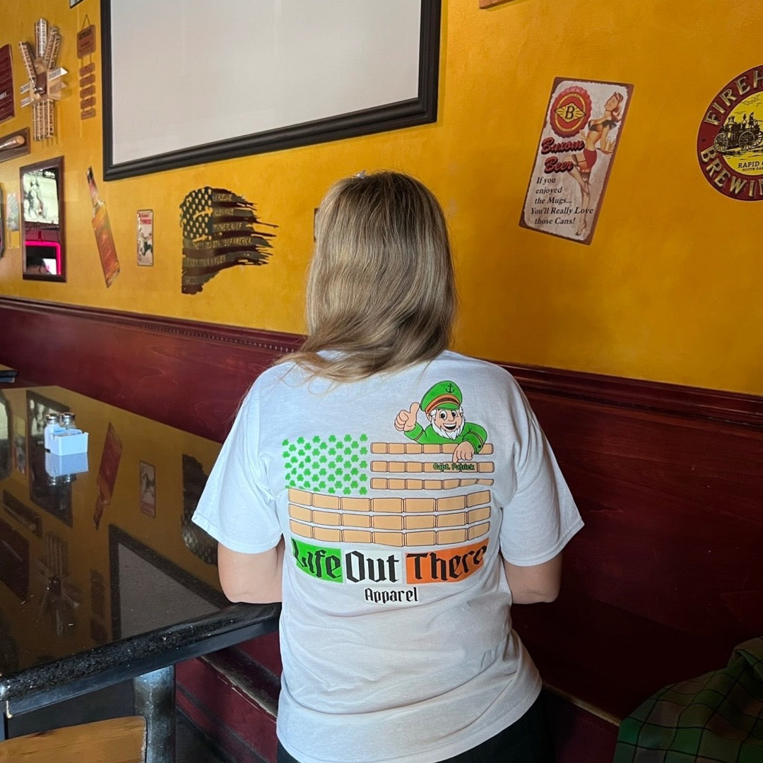 Life Out There Apparel “Capt. Patrick” Unisex tee shirt in white is a collaboration of an American flag with an Irish twist and a leprechaun boat Captain looking on. This is a photo,of a woman wearing the shirt at a bar / restaurant.  