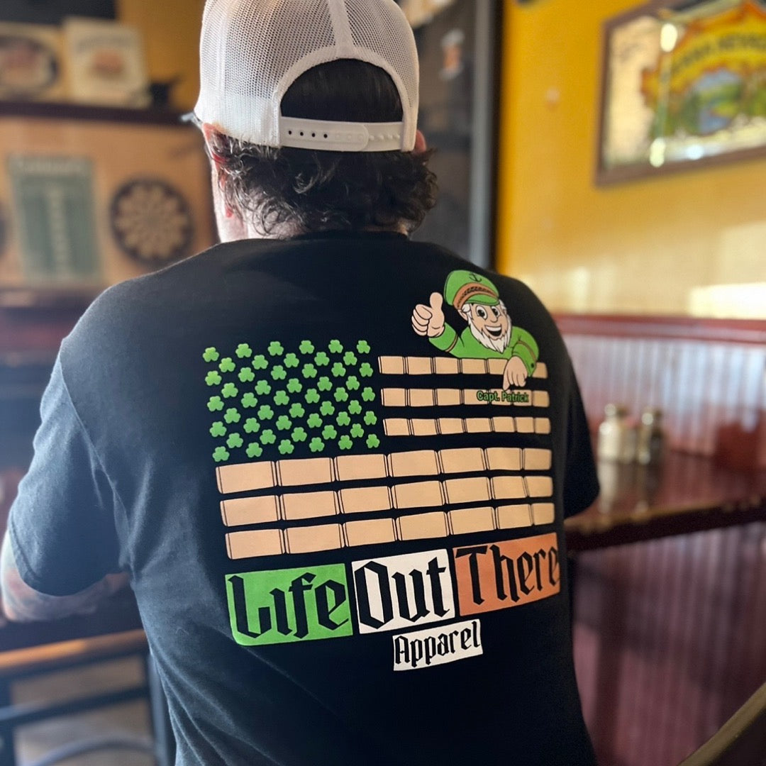 Life Out There Apparel “Capt. Patrick” Black Unisex tee shirt is a collaboration of an American flag with an Irish twist and a leprechaun boat Captain looking on.  Here a man is sitting in an Irish pub with the shirt on. 