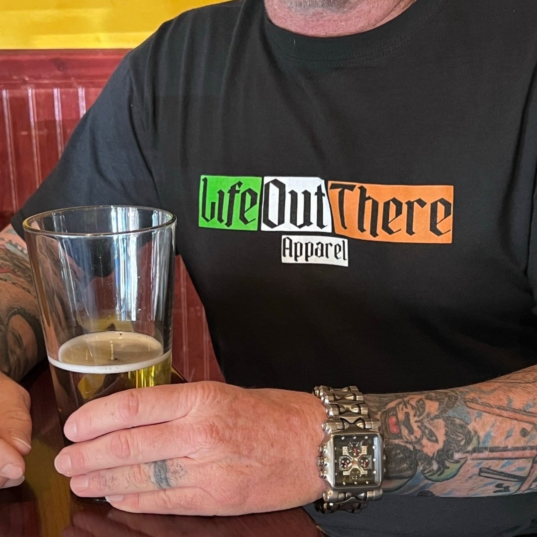 Life Out There Apparel “Capt. Patrick” black Unisex tee shirt is a collaboration of an American flag with an Irish twist and a leprechaun boat Captain looking on. This is the front of the shirt in the Irish flag colors. 